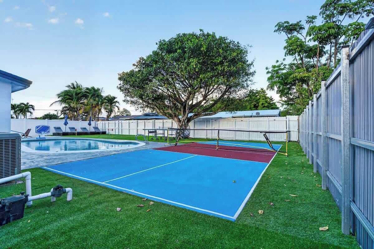 A backyard with a pool, artificial grass, a tennis court, and a large tree.