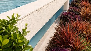 A swimming pool with a white marble wall, a blue tiled edge, and a variety of colorful plants. - Fully Furnished Duplex Apartment in Porto Seguro - Swimming Pool - 200m from the Beach (Porto Seguro)