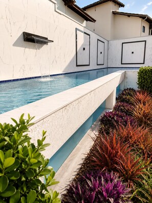 A swimming pool with a white marble wall, a blue tiled edge, and a variety of colorful plants. - Fully Furnished Duplex Apartment in Porto Seguro - Swimming Pool - 200m from the Beach (Porto Seguro)