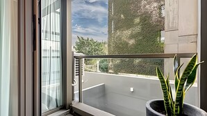 En balkon med en plantekasse, glasdør og udsigt til gade og grøntområde.