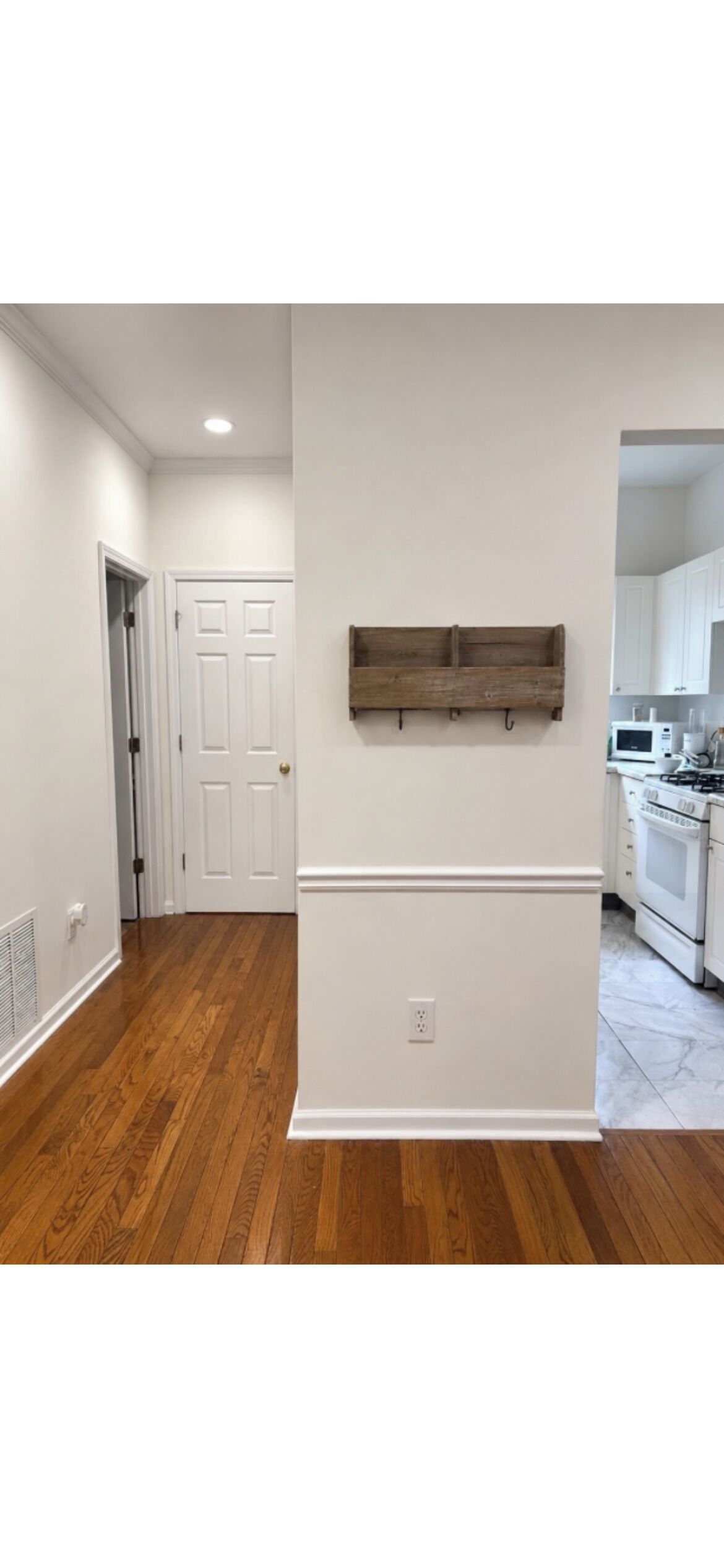 A hallway with wooden flooring, a white door, a wall-mounted wooden shelf, and a kitchen area with white cabinets and appliances.