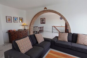 A living room with two sofas, a coffee table, a lamp, a dining area with chairs, and a staircase. - R1360 Pueblo Alhama 54 (Nerja)