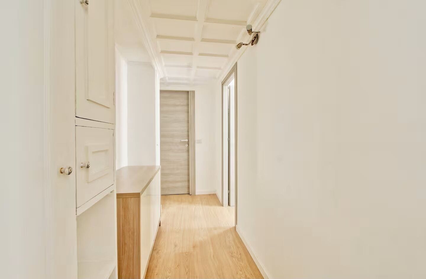 A narrow hallway with white walls and wooden flooring, leading to a door with a light switch.