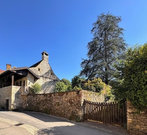 Ein Steinhaus mit einem kegelförmigen Dach, einem Holzzaun und einem großen Baum im Hintergrund.