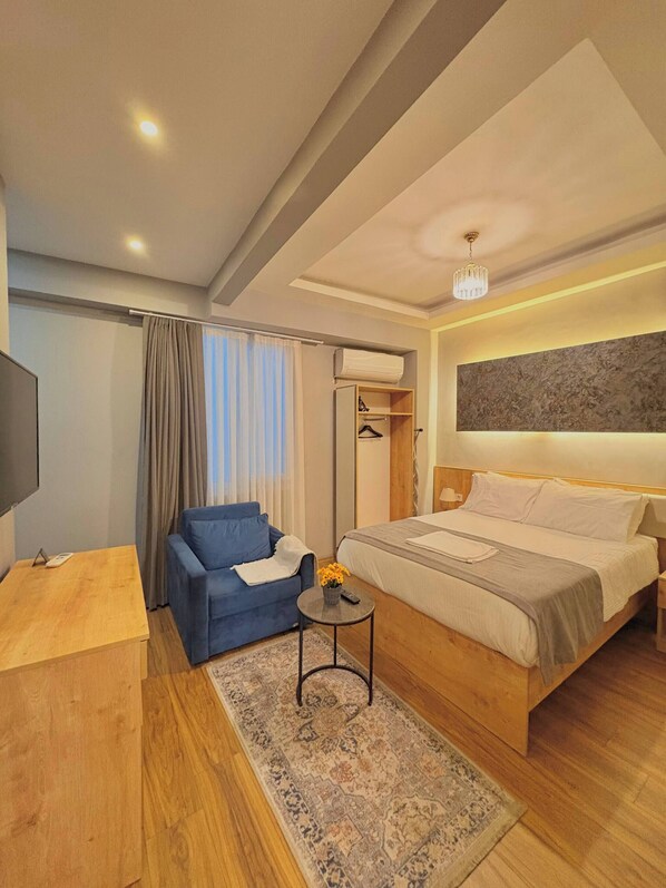 Ein Hotelzimmer mit einem Bett, einem blauen Sessel, einem kleinen Tisch mit einer Fernbedienung, einem Teppich und einem Fernseher auf einem Holzboden.