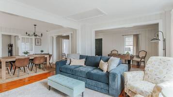 Un salón espacioso con un sofá azul, una zona de comedor con mesa y sillas de madera, y un sillón estampado.