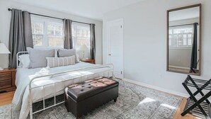 Un dormitorio con cama, mesitas de noche, espejo, ventana con cortinas y una alfombra estampada.