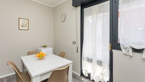 Een eetgedeelte met een witte tafel, beige stoelen, een schaal fruit, een klok en een raam met transparante gordijnen.