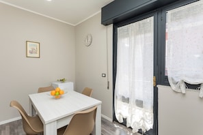 A dining area with a white table, beige chairs, a fruit bowl, a clock, and a window with sheer curtains.