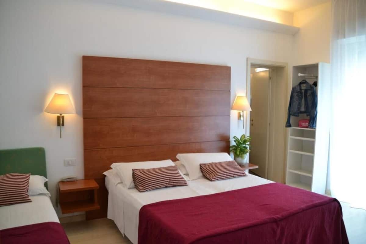 Habitación de hotel con un gran cabecero de madera, dos camas con almohadas a juego, lámparas de noche, una planta y un armario con ropa colgada.