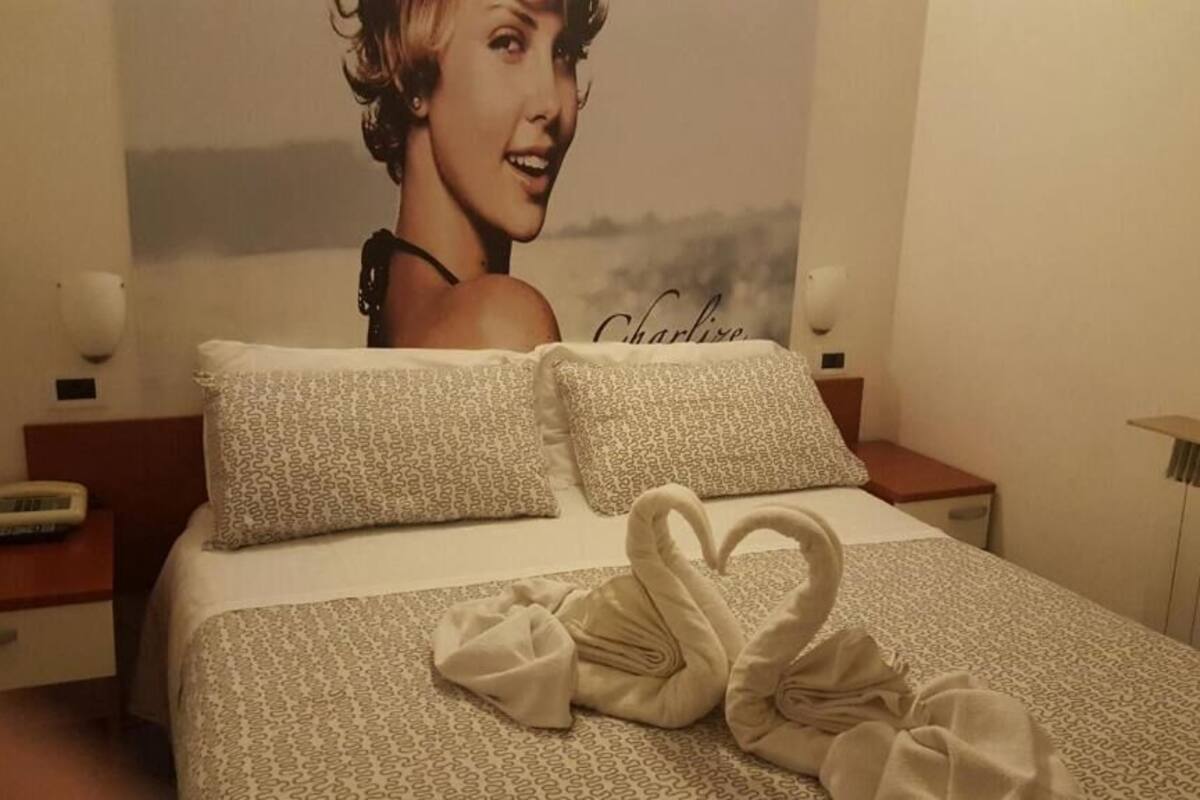 Una habitación de hotel con una cama, almohadas, una toalla con forma de cisne, un cartel en la pared y una mesita de noche con un teléfono.