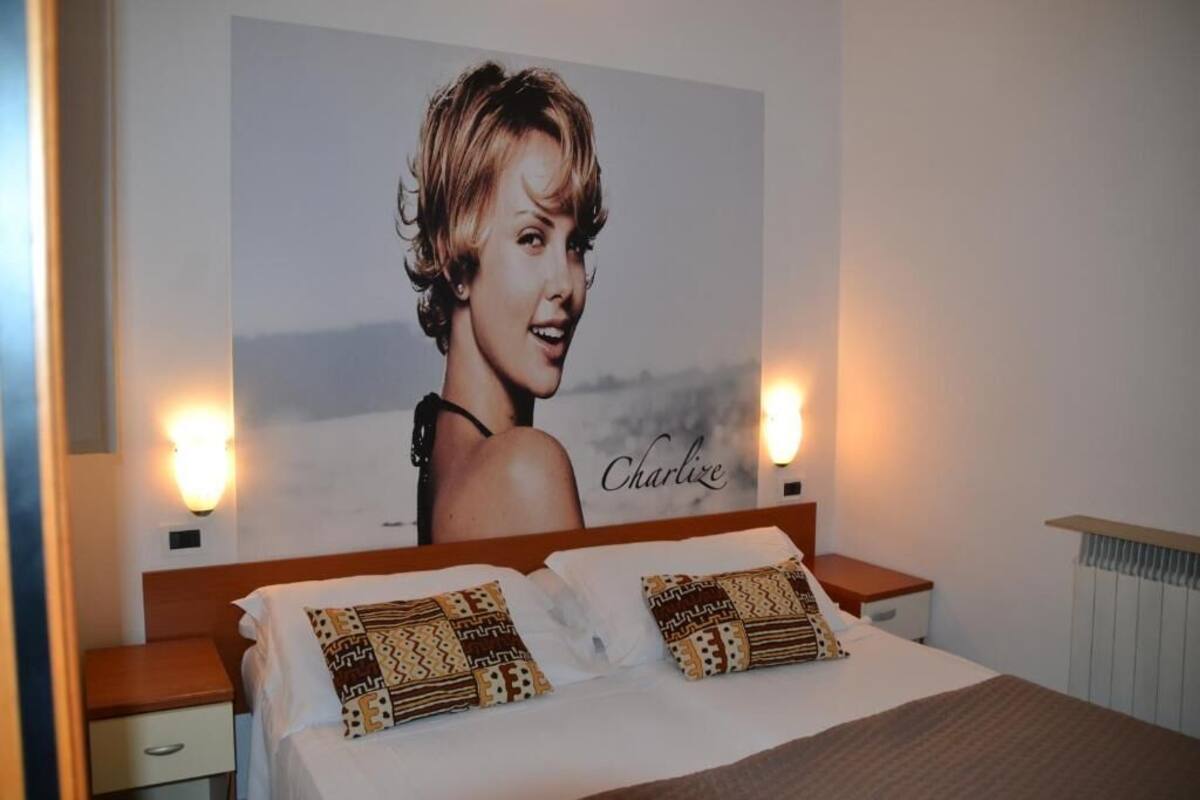 Habitación de hotel con una cama, dos mesitas de noche, una lámpara y un cartel en la pared, además de una ventana.