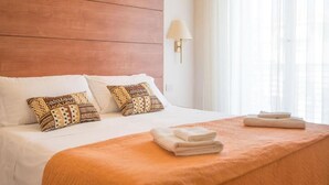 Ein Hotelzimmer mit Holz-Kopfteil, weißer Bettwäsche, gemusterten Kissen, einer beigen Tagesdecke, einem gefalteten Handtuch und einer Wandleuchte.