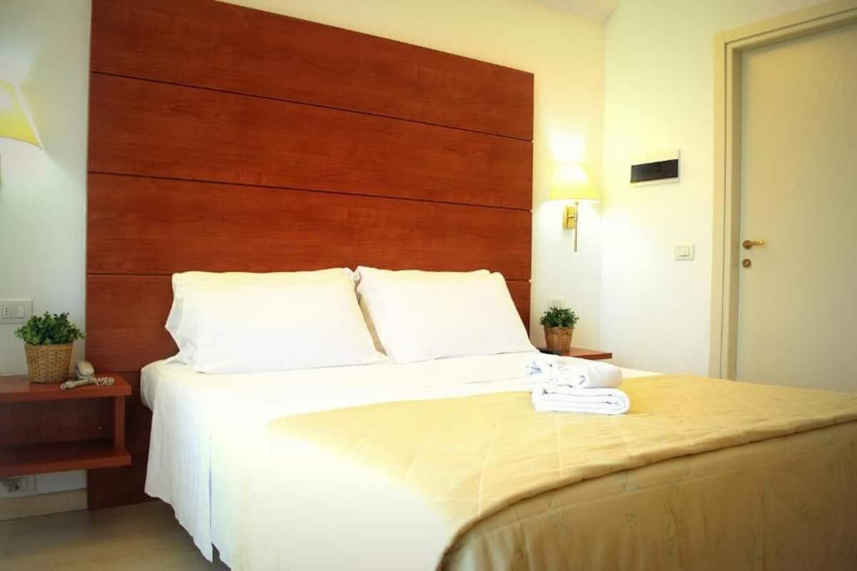Una habitación de hotel con cabecera de madera, una cama con ropa blanca, dos mesitas de noche con lámparas y una puerta.