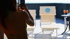 Eine Person macht ein Foto mit einem Smartphone, ein Balkon mit Stühlen und ein Tisch mit zwei Gläsern Getränken.