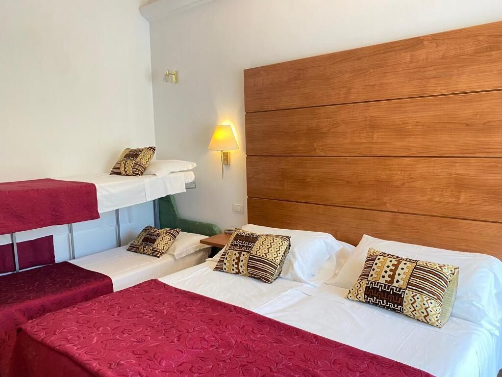 A hotel room with two beds, a wooden headboard, a bedside table, a lamp, and a red bedspread.