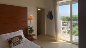Ein Hotelzimmer mit Bett, Nachttisch mit Lampe, Topfpflanze, Schiebetür zu einem Balkon und Blick ins Freie.
