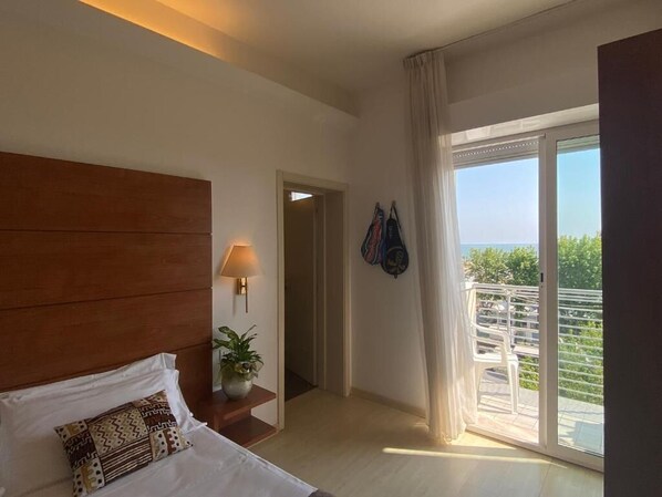 Habitación de hotel con cama, mesita de noche con lámpara, planta en maceta, vista al exterior a través de una puerta corrediza y balcón con barandilla.