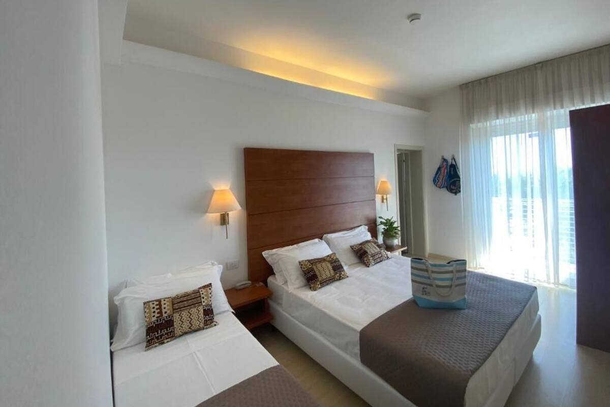 Habitación de hotel con una cama grande, una cama más pequeña, una mesita de noche, una lámpara, una ventana con cortinas y vistas a edificios.