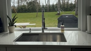 A kitchen with a stainless steel sink, a potted plant, and a view of a backyard with trees and a barbecue grill. - Peaceful and updated 4-bedroom house in East Quogue (East Quogue)
