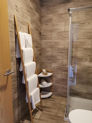A bathroom with a wooden wall, a glass shower enclosure, a wooden towel rack, and a small shelf with towels. - Atlantic Nature Beach House (BARRA DE MIRA)