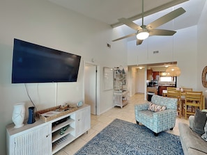 A living room with a flat-screen TV, a ceiling fan, a patterned rug, and a dining area with wooden furniture. - The Landing #104 (Gulf Shores)