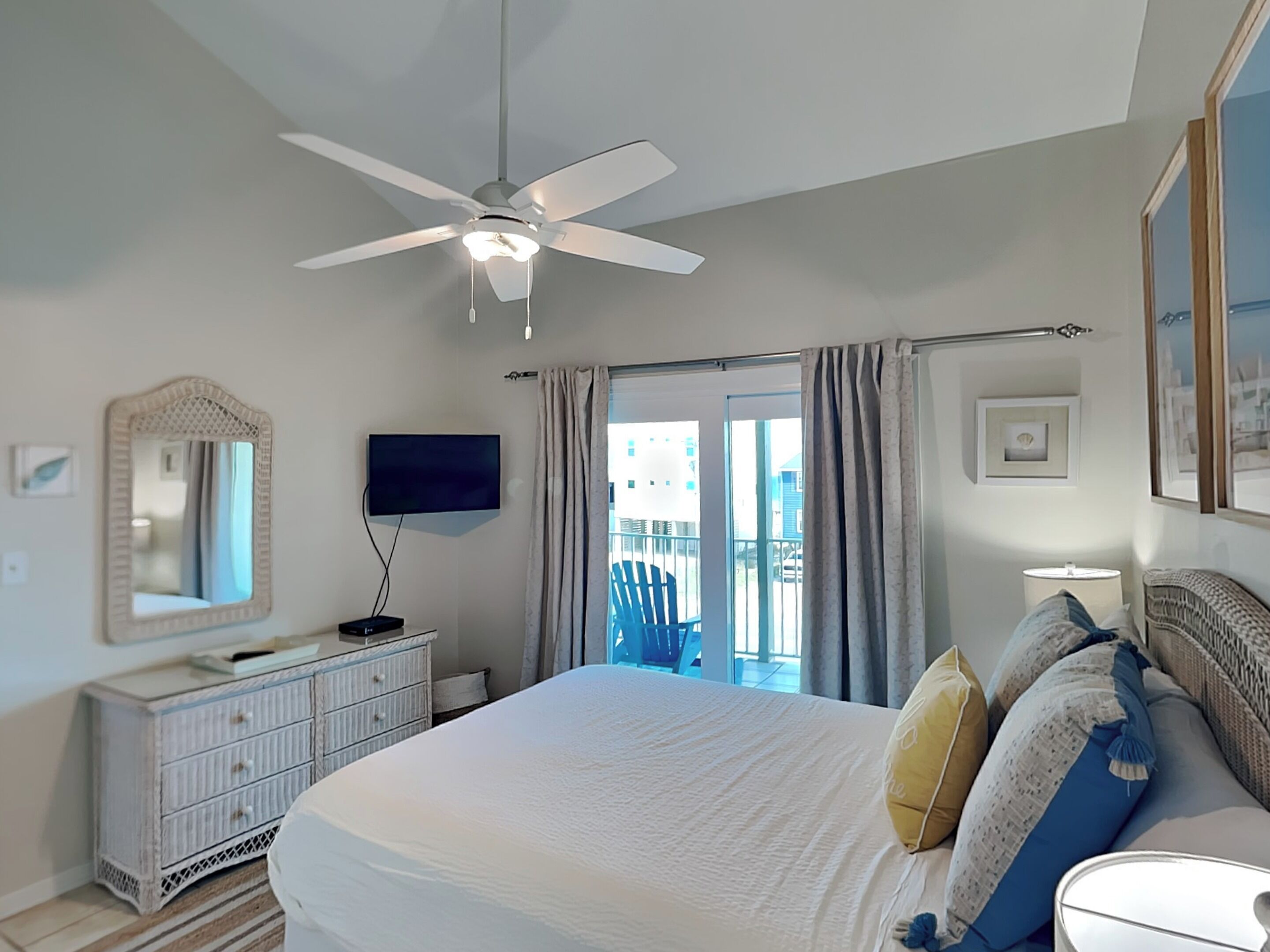 A bedroom with a bed, a ceiling fan, a TV, a dresser, a balcony with a chair, and a window with curtains.