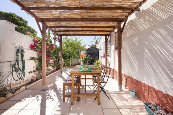 Eine überdachte Terrasse mit Holztisch und Bänken, umgeben von einer weißen Mauer mit rotem Sockel, und Blumentöpfen.
