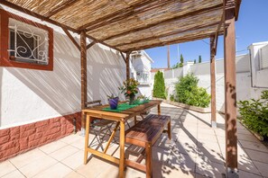Eine überdachte Terrasse mit einem Holztisch und einer Bank, ein Fenster mit schmiedeeisernen Verzierungen und eine weiße Wand mit rotem Fußbrett.