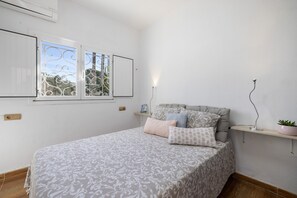 Ein Schlafzimmer mit Bett, einem Fenster mit Metallgitter, einem Nachttisch mit Pflanze und einem an der Wand befestigten Regal.