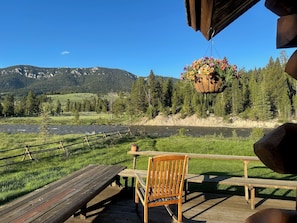 Eine Holzterrasse mit einer Bank und einem Tisch, mit Blick auf einen Fluss und bewaldete Berge.
