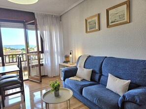 Un salon avec un canapé bleu, une table à manger avec des chaises, une petite table ronde avec une plante, et un balcon avec vue.
