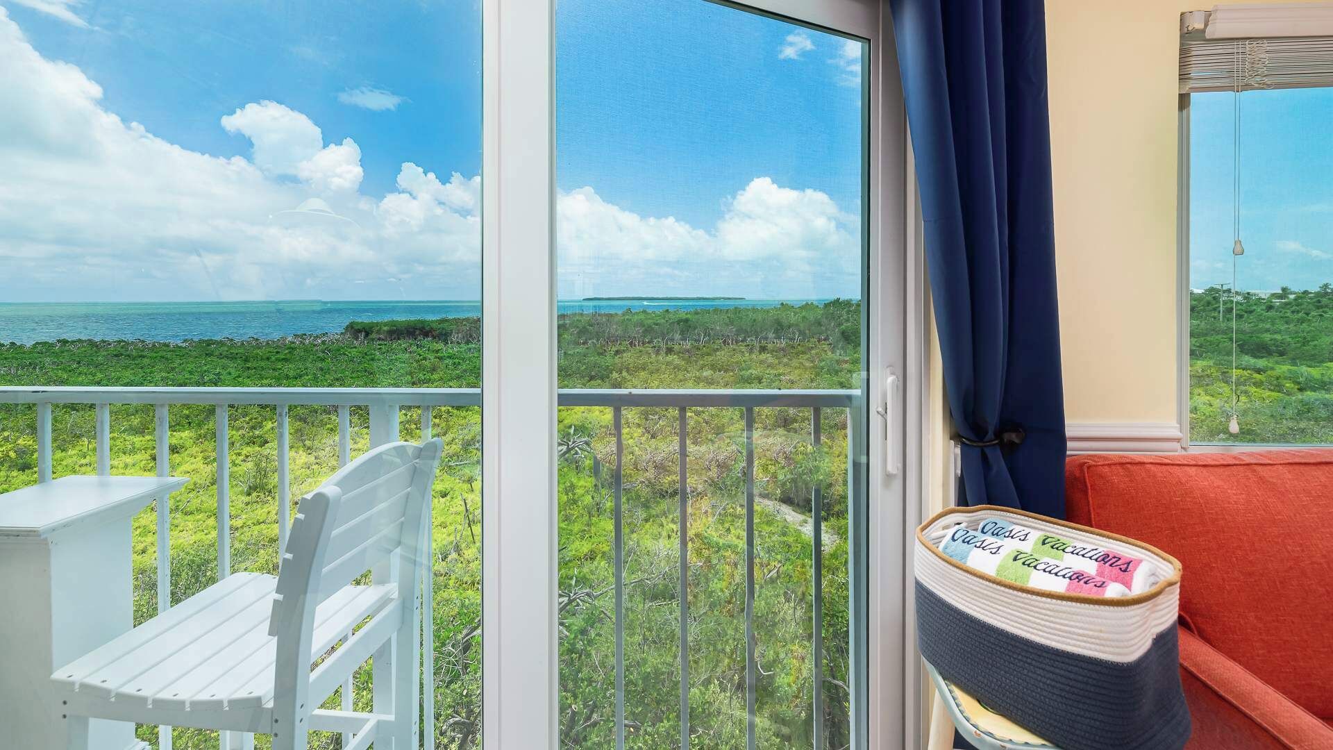 A balcony with a white chair, a small table, and a view of the ocean and greenery.