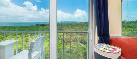 A balcony with a white chair, a small table, and a view of the ocean and greenery.