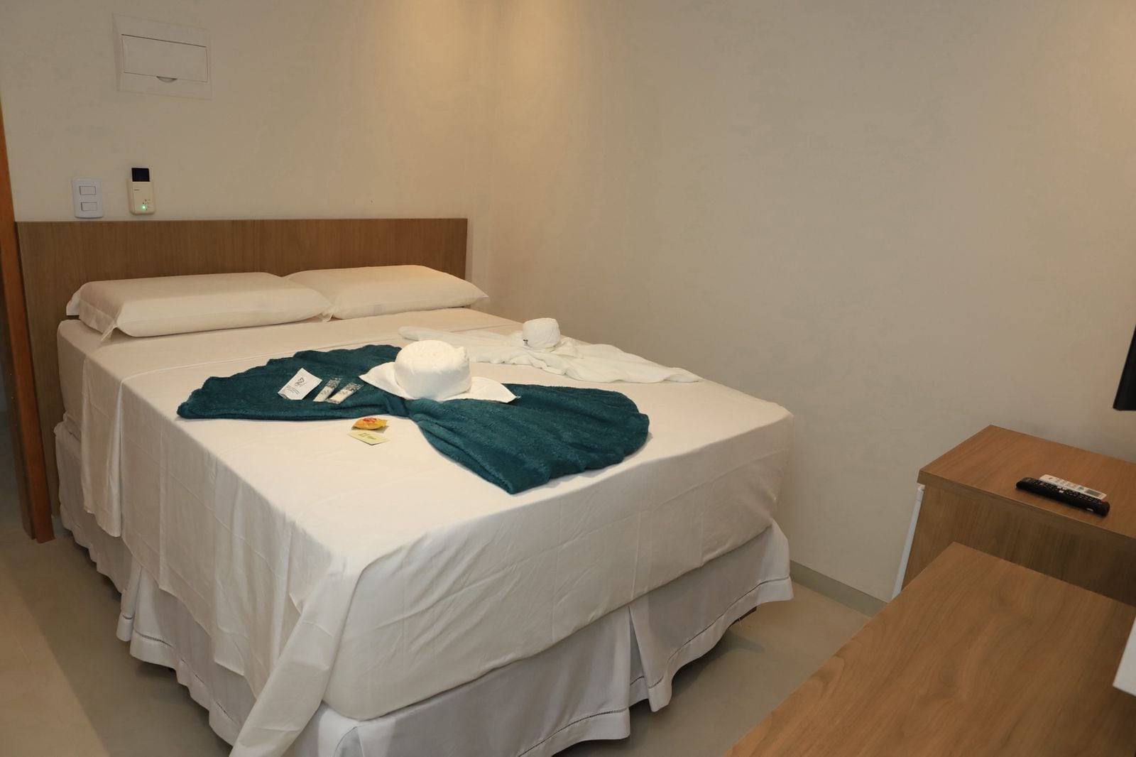Quarto de hotel com cama, mesa de cabeceira com controle remoto e termostato fixado na parede.