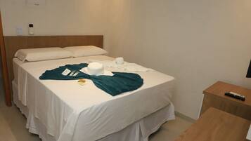 Quarto de hotel com cama, mesa de cabeceira com controle remoto e termostato fixado na parede.
