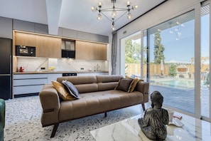 A modern kitchen with a built-in oven, a dining area with a sofa and a statue, and a large window with a view of a swimming pool. - Nightingale Alexander Villa - private pool (Paleokastritsa)