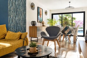 Ein modernes Wohnzimmer mit einer Couch, einem runden Couchtisch mit einer Pflanze, einem grauen Sessel und einem Essbereich mit Tisch und Stühlen.