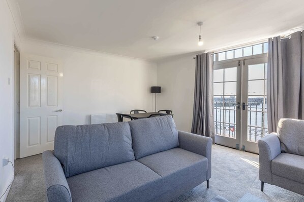 Un salon avec un canapé gris, un fauteuil gris, une table à manger avec des chaises, un lampadaire et une vue à travers une grande fenêtre avec des rideaux.