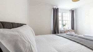 A hotel room with a bed, a bedside table with a lamp, and a window with curtains. - Modern 2BR Sea View Flat on the Harbour (Whitehaven)