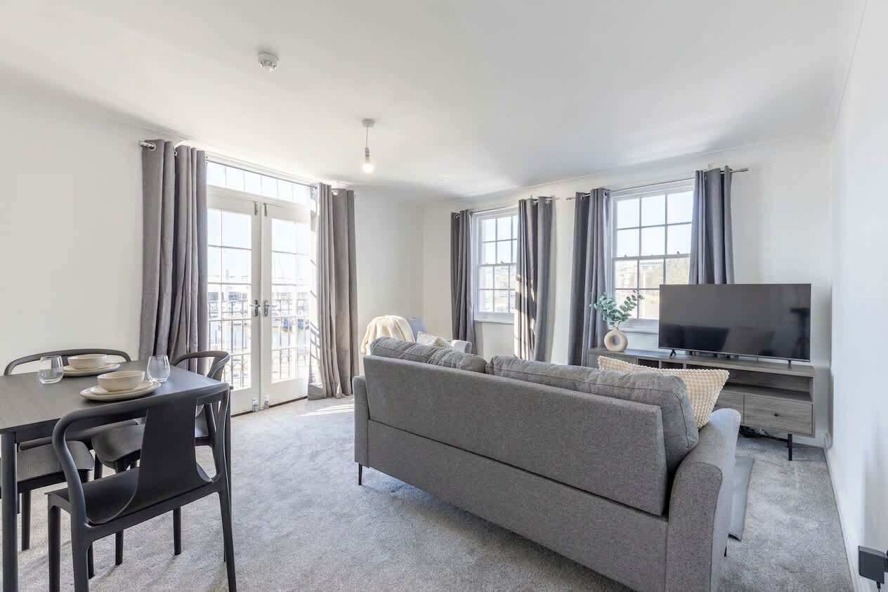 A modern living room with a sofa, a dining table with chairs, a flat-screen TV, and large windows with curtains.