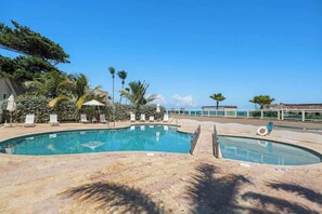Ein Swimmingpool mit Liegestühlen und Sonnenschirmen, umgeben von Palmen und blauem Himmel.