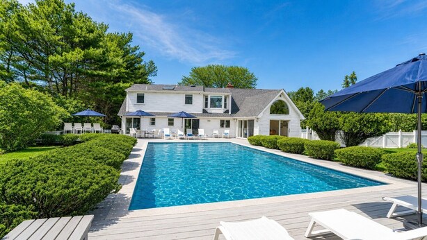 Ein Schwimmbecken mit Liegestühlen, ein Haus mit Veranda und ein weißer Zaun.