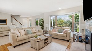 Ein geräumiges Wohnzimmer mit zwei beigen Sofas, einem Couchtisch und großen Fenstern mit Blick auf Grünflächen.