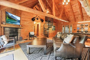 A log cabin interior with a stone fireplace, leather sofa, wooden ceiling beams, a spiral staircase, and a television mounted on the wall. - The Johnny: Sleeps 16 w/ Brand-New Pool, Hot Tub, Firepit & Private Pond (Laurelville)