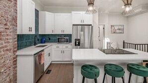 Una cucina moderna con mobili bianchi, un frigorifero in acciaio inossidabile, un forno a microonde e una macchina per il caffè. Sono presenti tre sgabelli da bar verdi e un lampadario.