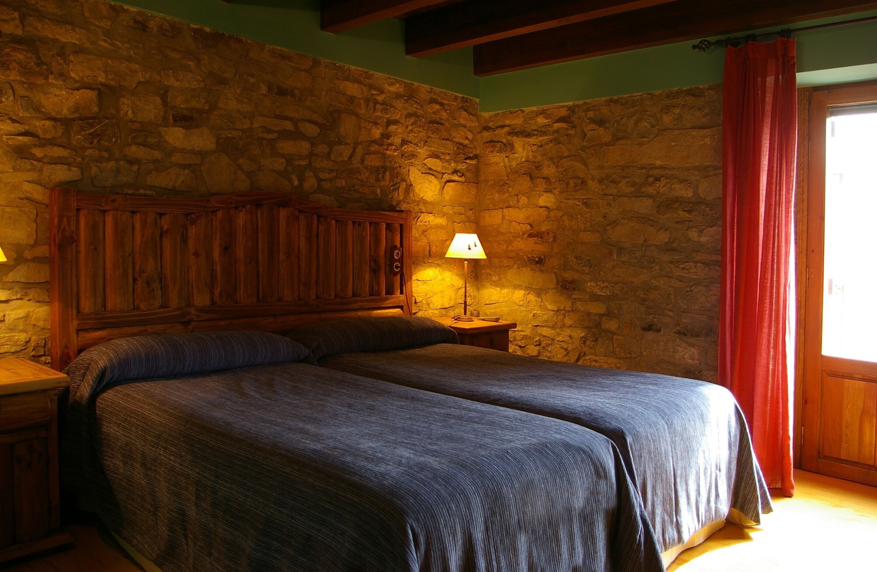 Ein Zimmer mit einer Steinswand, einem hölzernen Bett mit zwei Kissen, einem Nachttisch mit einer Lampe und einem roten Vorhang.