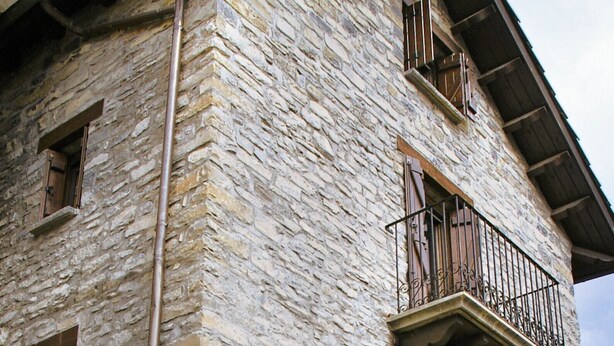 Un edificio de piedra con una puerta de madera, un balcón con barandilla de metal y una ventana con persiana de madera.