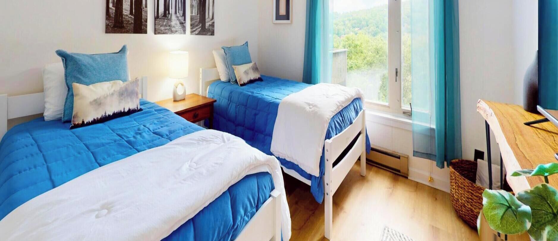 A bedroom with two beds, a bedside table with a lamp, a window with curtains, and a view of greenery outside.