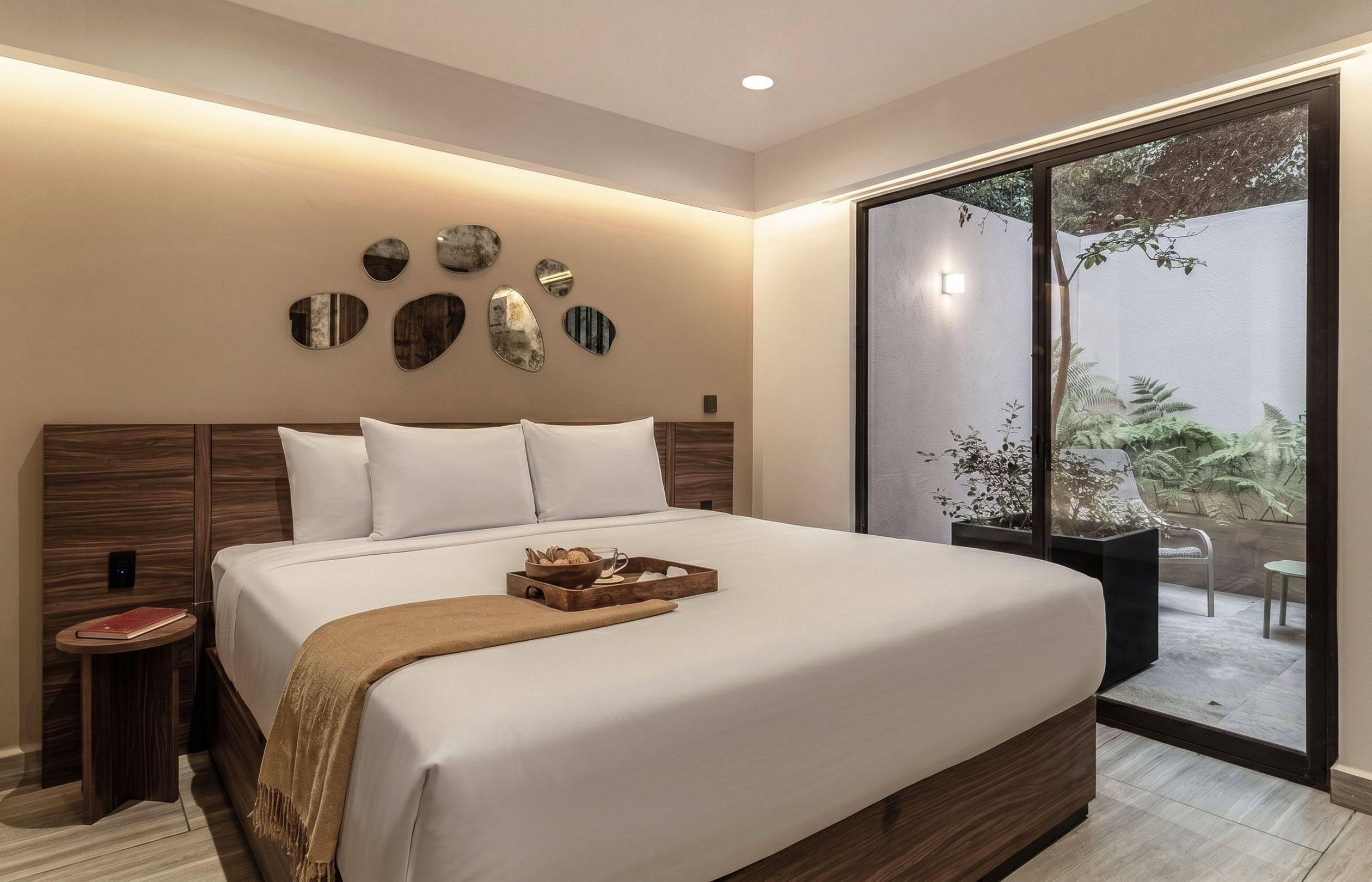 A modern bedroom with a large bed, bedside tables, decorative wall art, and a sliding glass door leading to an outdoor area with plants.